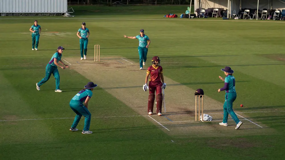Fielders in coloured women's team kits gather near the pitch between overs while a bowler adjusts her run-up marker on a sunlit outfield